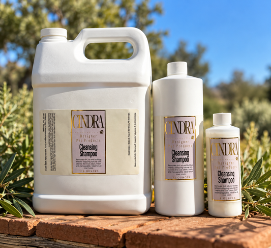 Three white bottles of Cindra Natural Products cleansing shampoo in different sizes, displayed outdoors on a brick surface with greenery and blue sky in the background.