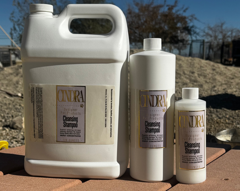 Three white containers of Cindra Cleansing Shampoo in different sizes arranged on a terracotta tile surface outdoors under a blue sky.